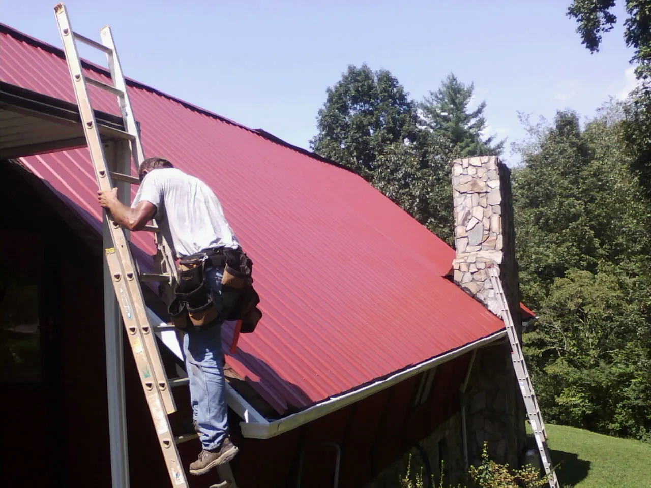 Roofer on a ladder beside a red metal roof and stone chimney — Soffit & Fascia Repair by Roofing Brothers in Rutland